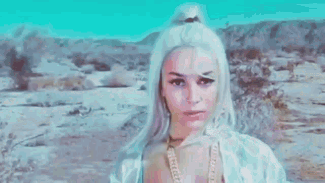 a woman with purple hair is standing in the desert wearing a white shirt and a necklace .