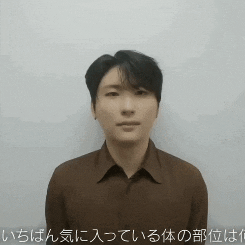 a man wearing a brown shirt is standing in front of a white wall with foreign writing on it