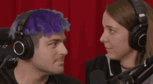 a man with purple hair and a woman wearing headphones are looking at each other in front of a red curtain .