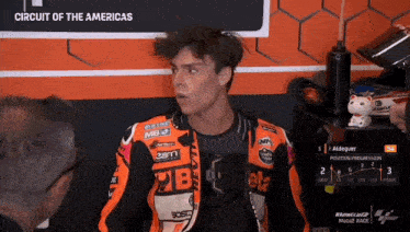 a man in an orange and black motorcycle jacket is talking to another man