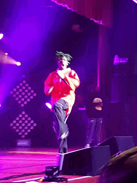 a person on a stage with purple lights behind them and a man in a black shirt with the letter m on it