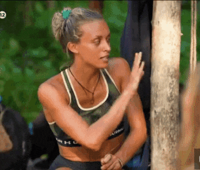 a woman in a sports bra is standing next to a tree and waving .
