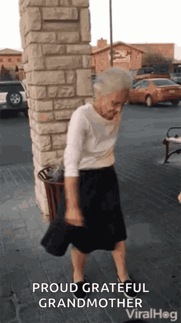 an elderly woman is standing in front of a brick wall and a trash can .