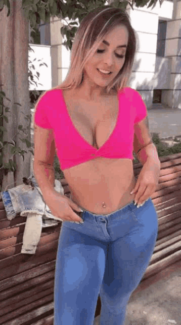 a woman wearing a pink crop top and blue jeans is standing next to a bench .
