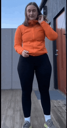 a woman wearing an orange jacket and black leggings is standing on a wooden deck