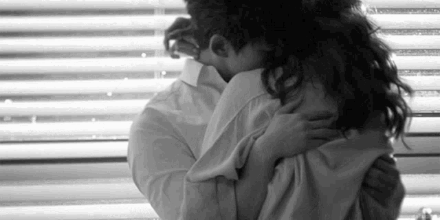 a black and white photo of a man and woman hugging each other in front of a window .