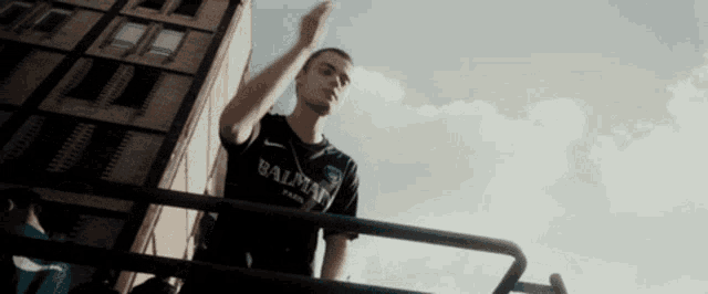 a man wearing a black balmain shirt is standing on a balcony