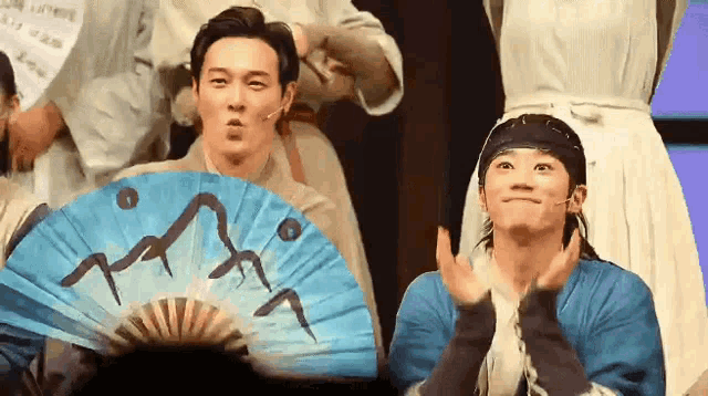 a man is holding a blue fan and making a funny face while another man applauds .