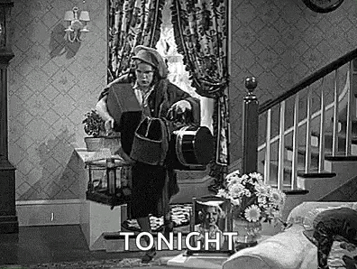 a black and white photo of a woman standing in a living room holding a dog .