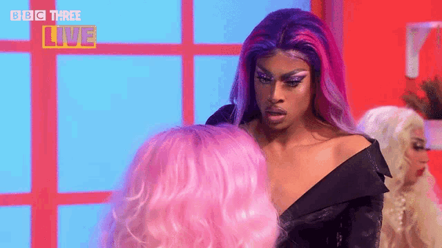 a drag queen with purple and pink hair is standing next to a pink wig .