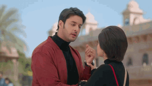 a man in a red jacket and black turtleneck talks to a woman