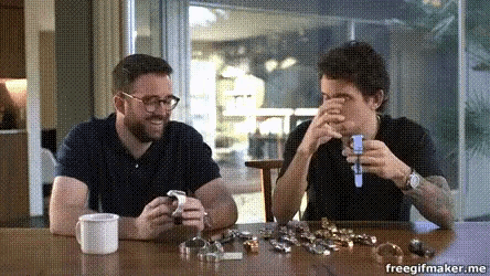 two men are sitting at a table looking at watches and the words freegifmaker.me are on the bottom right