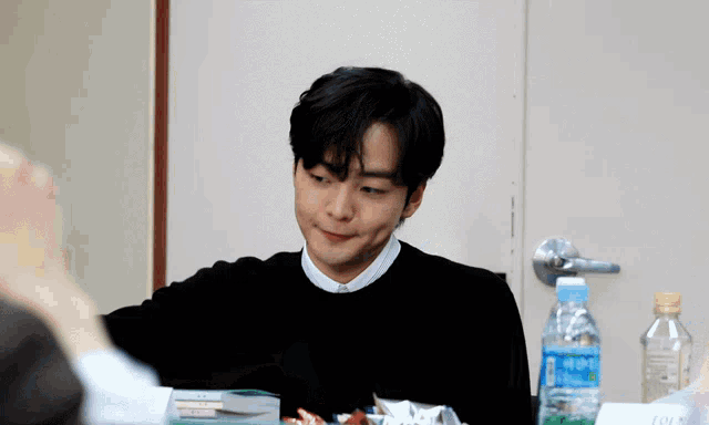 a man in a black sweater sits at a table with a bottle of water in front of him