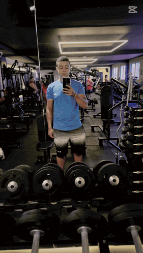 a man in a blue shirt takes a selfie in a gym with the number 18 on the ceiling