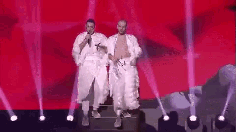 two men are singing into microphones on a stage while wearing white robes .
