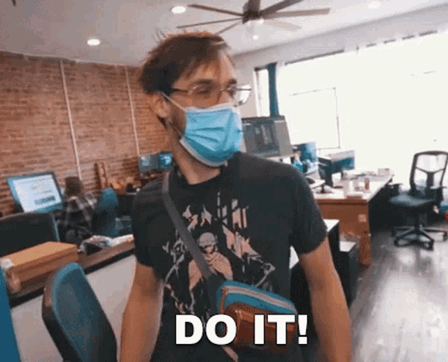 a man wearing a mask and a t-shirt is standing in an office and saying `` do it '' .