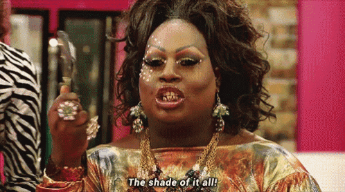 a drag queen says " the shade of it all " while holding a mirror