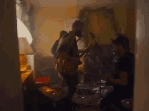 a man is playing a guitar in a living room while another man plays drums .