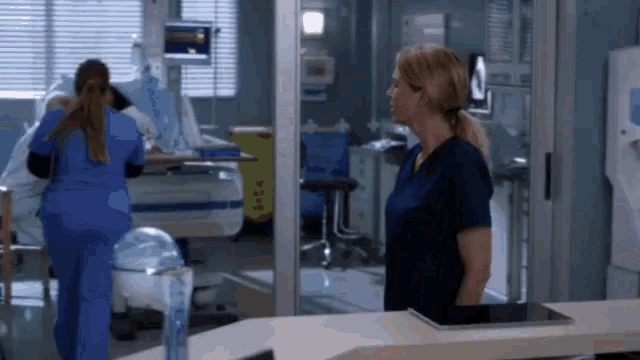 a woman in a blue scrub is standing in a hospital room next to a patient in a hospital bed .