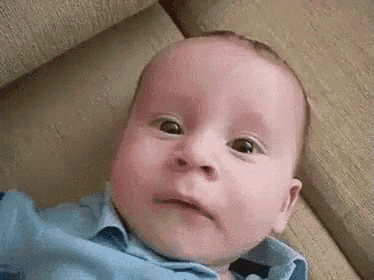 a baby in a blue shirt is laying on a couch and making a funny face .