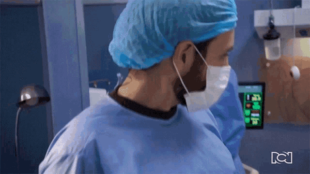 a surgeon wearing a mask and a blue cap stands in front of a monitor that says ' a ' on it