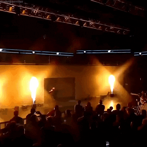 a man stands on a stage with flames coming out of the ceiling and a crowd watching