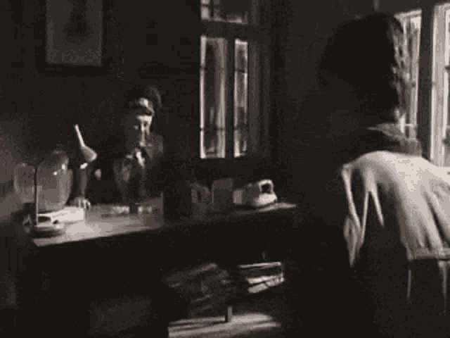 a black and white photo of a man standing in front of a desk talking on a phone .