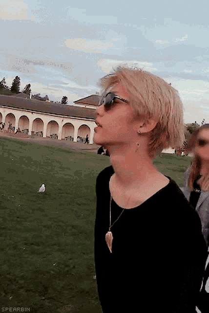 a man wearing sunglasses and a necklace stands in a field with a building in the background and the words spearbin below him