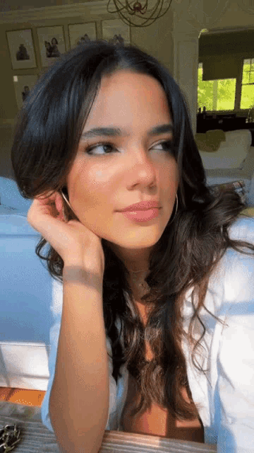 a woman with long dark hair is sitting in a living room with her hand on her chin