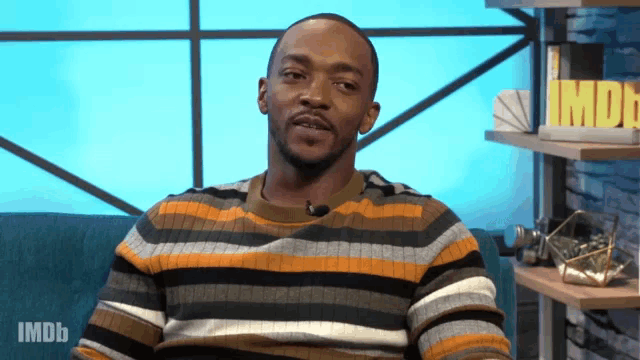 a man in a striped sweater sits on a blue couch with imdb written on the bottom right