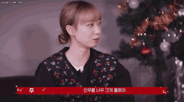 a woman is sitting in front of a christmas tree with a red sign that says honeycam.org