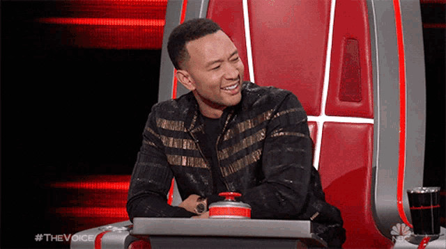 a man in a black jacket is sitting in a chair with a red button in front of him .