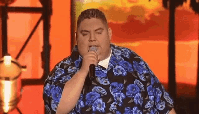 a fat man is singing into a microphone while wearing a blue floral shirt .