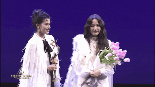 a woman holding a trophy and a bouquet of flowers stands next to another woman holding a bouquet of pink tulips