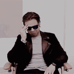 a man wearing sunglasses and a leather jacket is sitting in a chair and waving his hand .