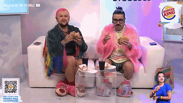 two men are sitting on a couch eating hamburgers with a burger king logo behind them