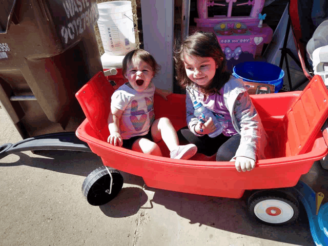 two little girls are sitting in a red wagon in front of a waste bin