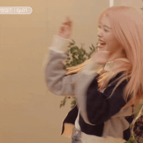 a woman with pink hair is standing in a room with her arms in the air and smiling .