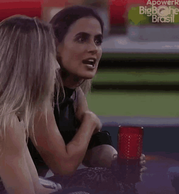 two women sitting next to each other with a red glass in front of a big brother brasil logo