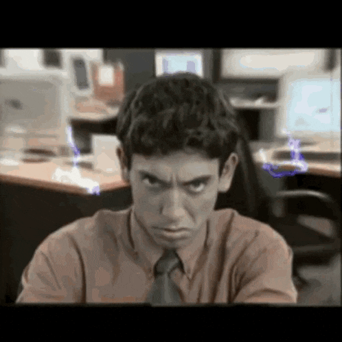 a man in a brown shirt and tie is sitting at a desk in front of a computer monitor .