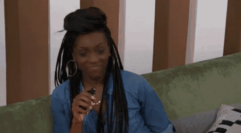 a woman with braids is sitting on a couch holding a cell phone and smiling .