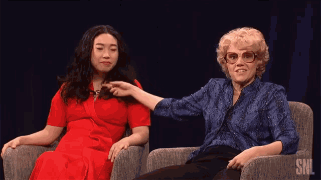 two women are sitting in chairs and one is pointing at the other 's hair .