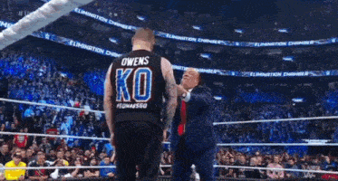 a man in a suit and tie is standing next to a man in a wrestling ring wearing a ko shirt .