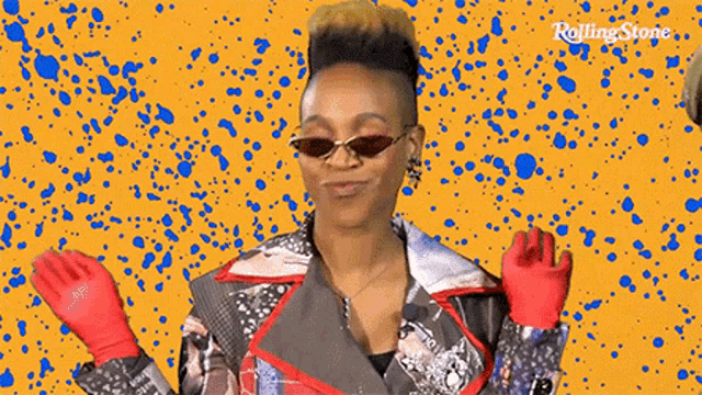 a woman wearing sunglasses and red gloves is standing in front of a rolling stone logo .