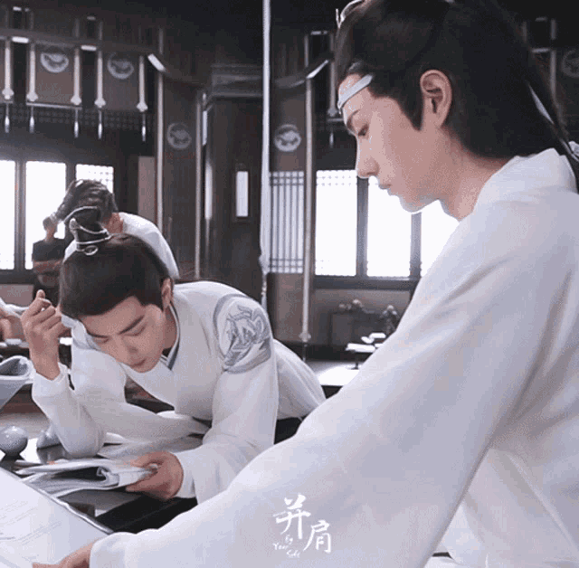 a man in a white shirt is looking at a book while another man looks on
