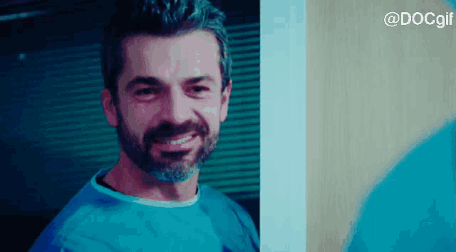 a man with a beard wearing a blue scrub top smiles in front of a door