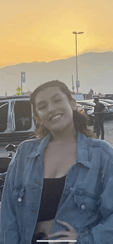 a woman in a denim jacket is smiling in a parking lot with mountains in the background .