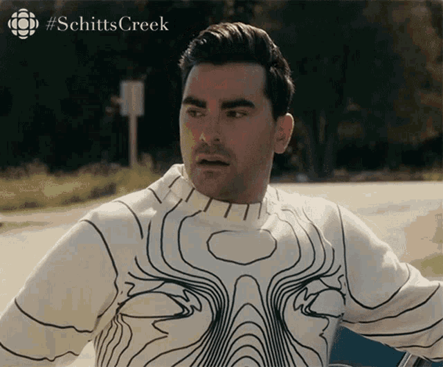a man wearing a white sweater with black lines and the word schitts creek on the bottom right