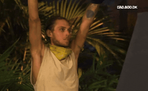 a man in a white tank top with a yellow bandana around his neck stands with his arms in the air in front of a palm tree