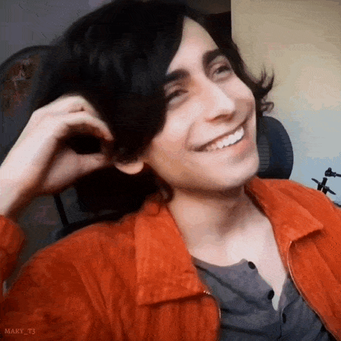a young man with long black hair is smiling while wearing an orange jacket and a grey shirt .
